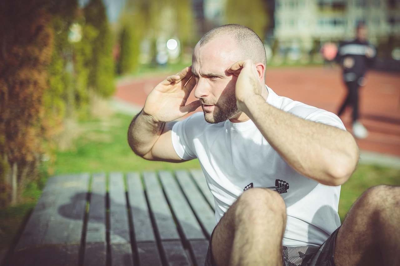 A man sitting on a wooden bench in a park. He is wearing a white t-shirt with a black logo on it and shorts. He has a bald he...
