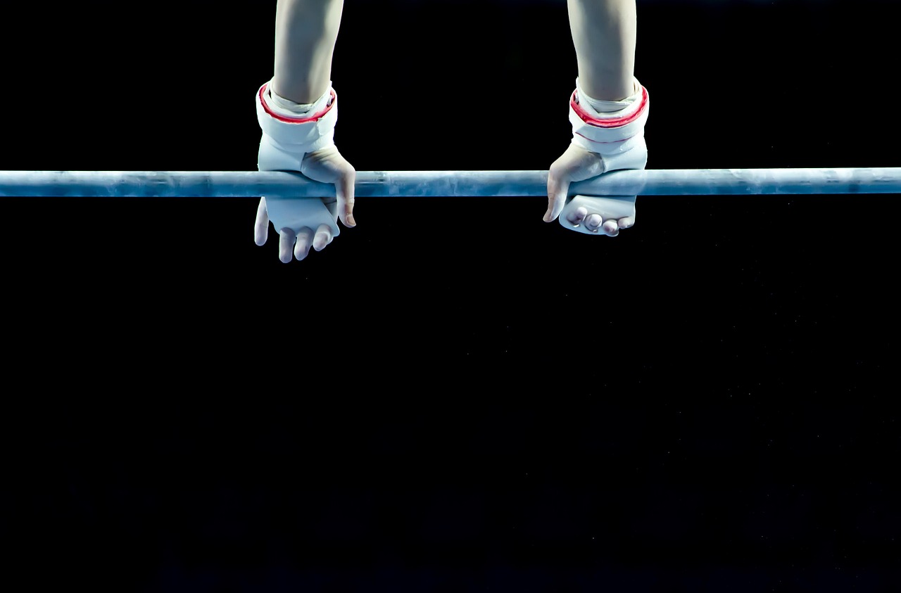 A pair of hands holding onto a horizontal bar. The hands are wearing white gloves with red stripes on the wrists and fingers....