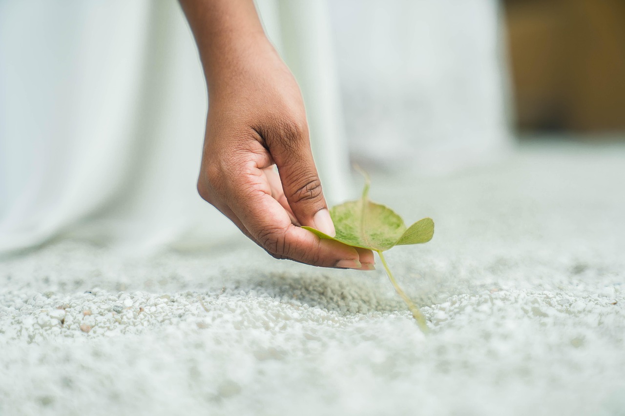 A person's hand holding a small green leaf. The hand is positioned in a way that the fingers are gently cradling the leaf, with the thumb and index finger on the left side of the image. The leaf appears to be fresh and healthy, with a few small veins running through it. The background is blurred, but it seems to be a white surface with small pebbles scattered around. The overall mood of the photo is peaceful and serene.