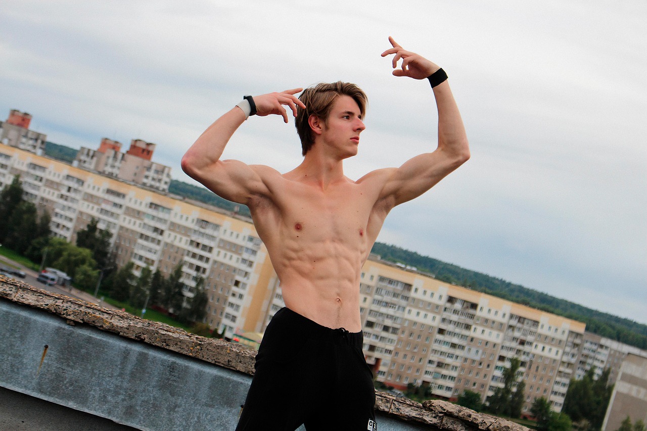 A young man standing on a rooftop with his arms raised above his head. He is shirtless and wearing black shorts. He has short...