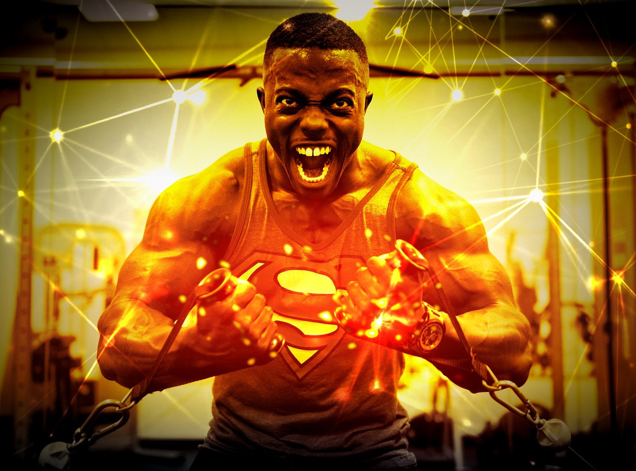 A young African-American man in a gym, wearing a Superman tank top. He is holding a pair of dumbbells in his hands and appear...