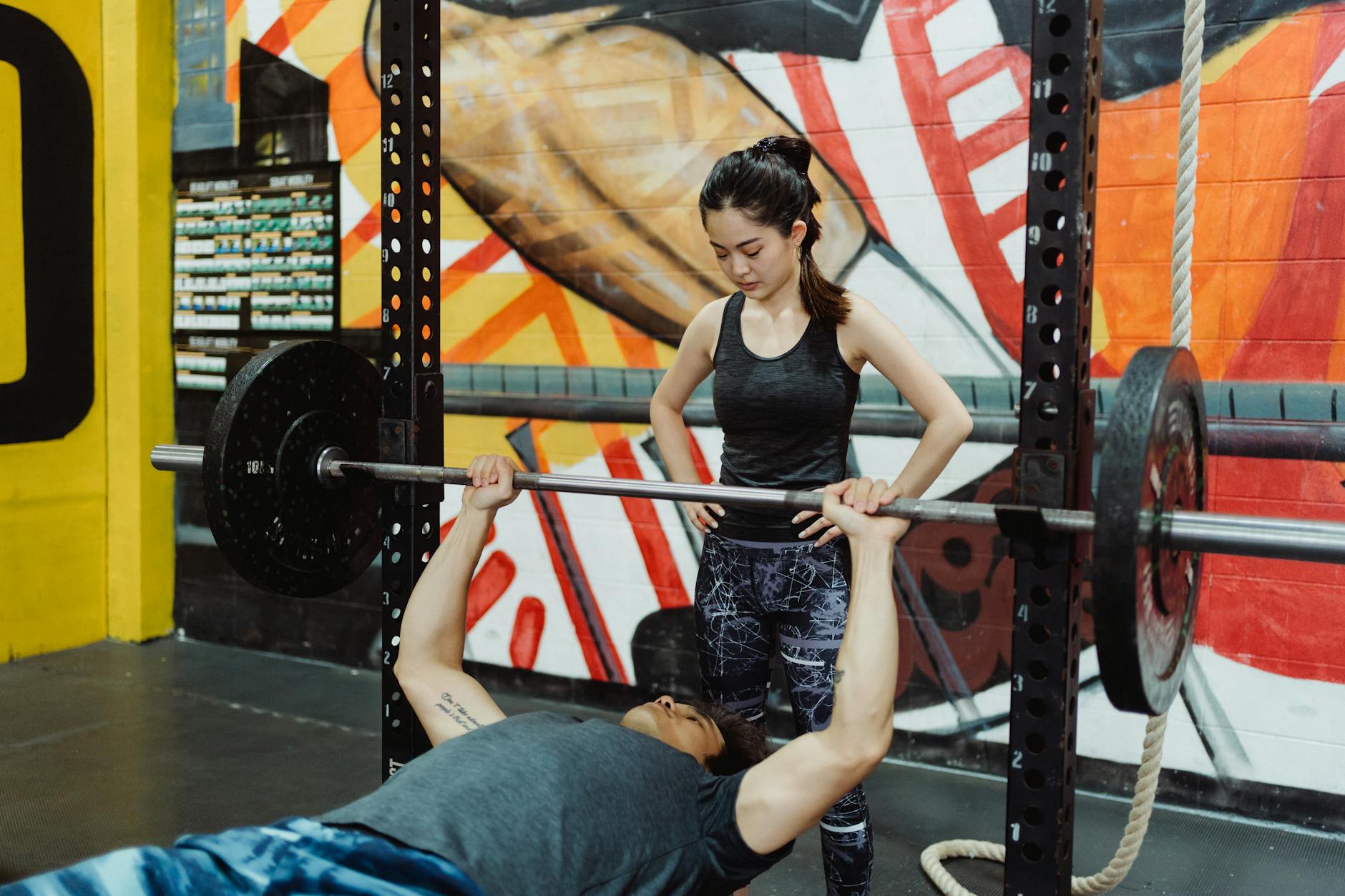 A man and a woman in a gym. The man is lying on his back with his arms stretched out in front of him, lifting a barbell over ...