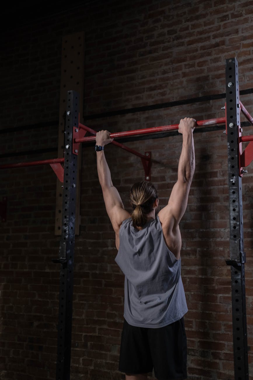 A man performing a pull-up exercise on a horizontal bar in a gym. He is wearing a grey tank top and black shorts and is holdi...