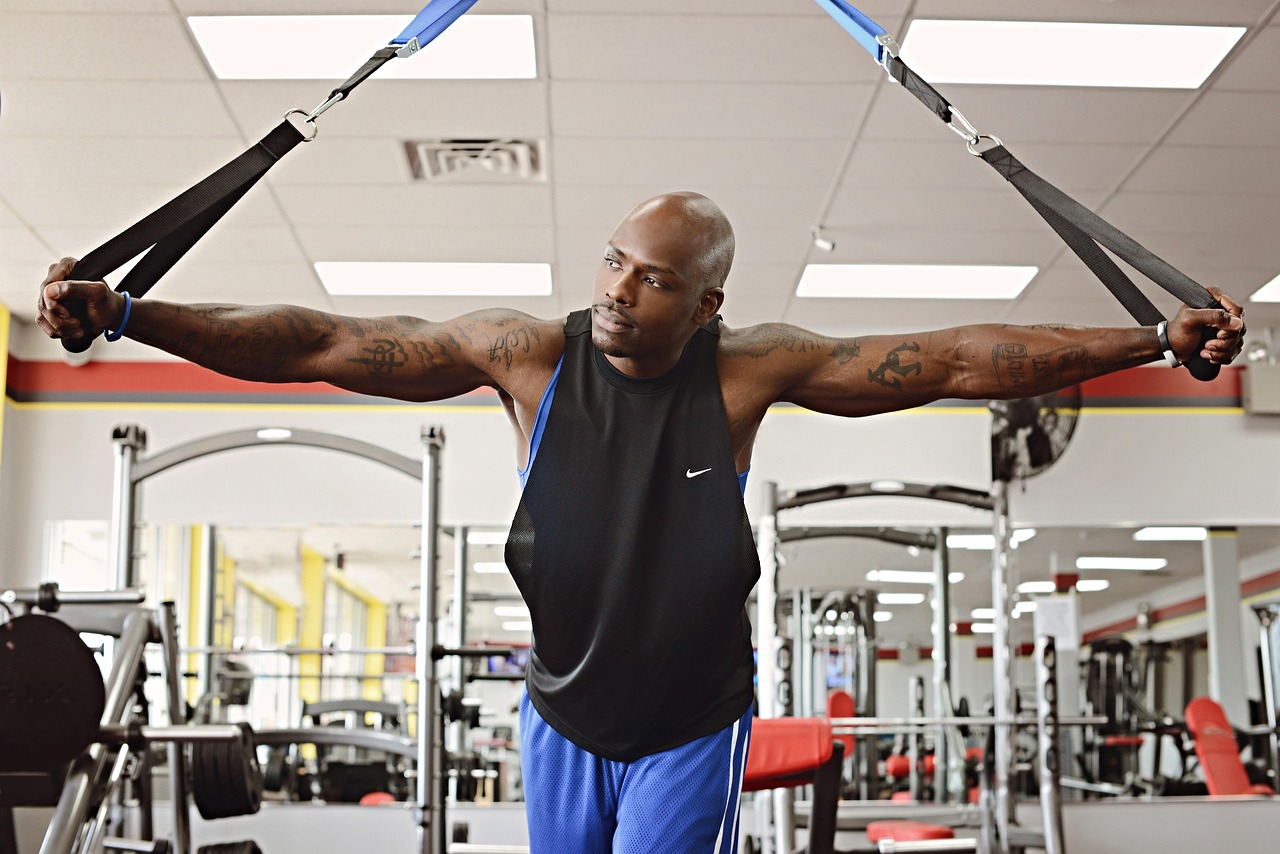 A man in a gym performing a TRX exercise. He is wearing a black tank top and blue shorts and has multiple tattoos on his arms and chest. The man is holding the TRX straps above his head with both hands and is in the middle of a pull-up exercise. In the background, there are various gym equipment and mirrors on the walls. He appears to be focused and determined as he performs the exercise.