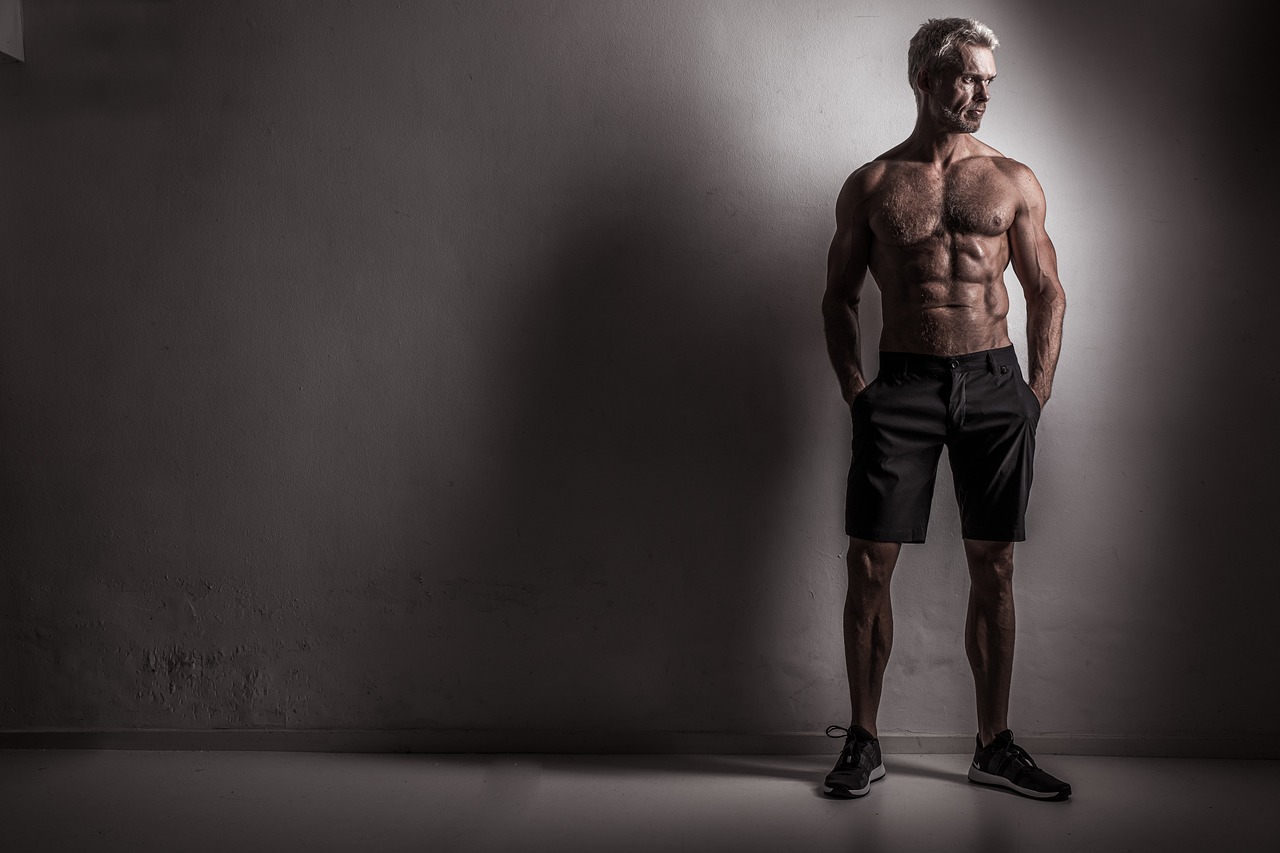 A black and white photograph of a muscular man standing in an empty room. He is shirtless and wearing black shorts and sneake...