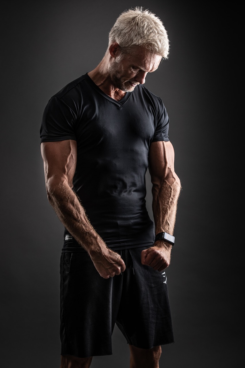A portrait of a middle-aged man standing in a dark room. He is wearing a black t-shirt and black shorts. His body is muscular...