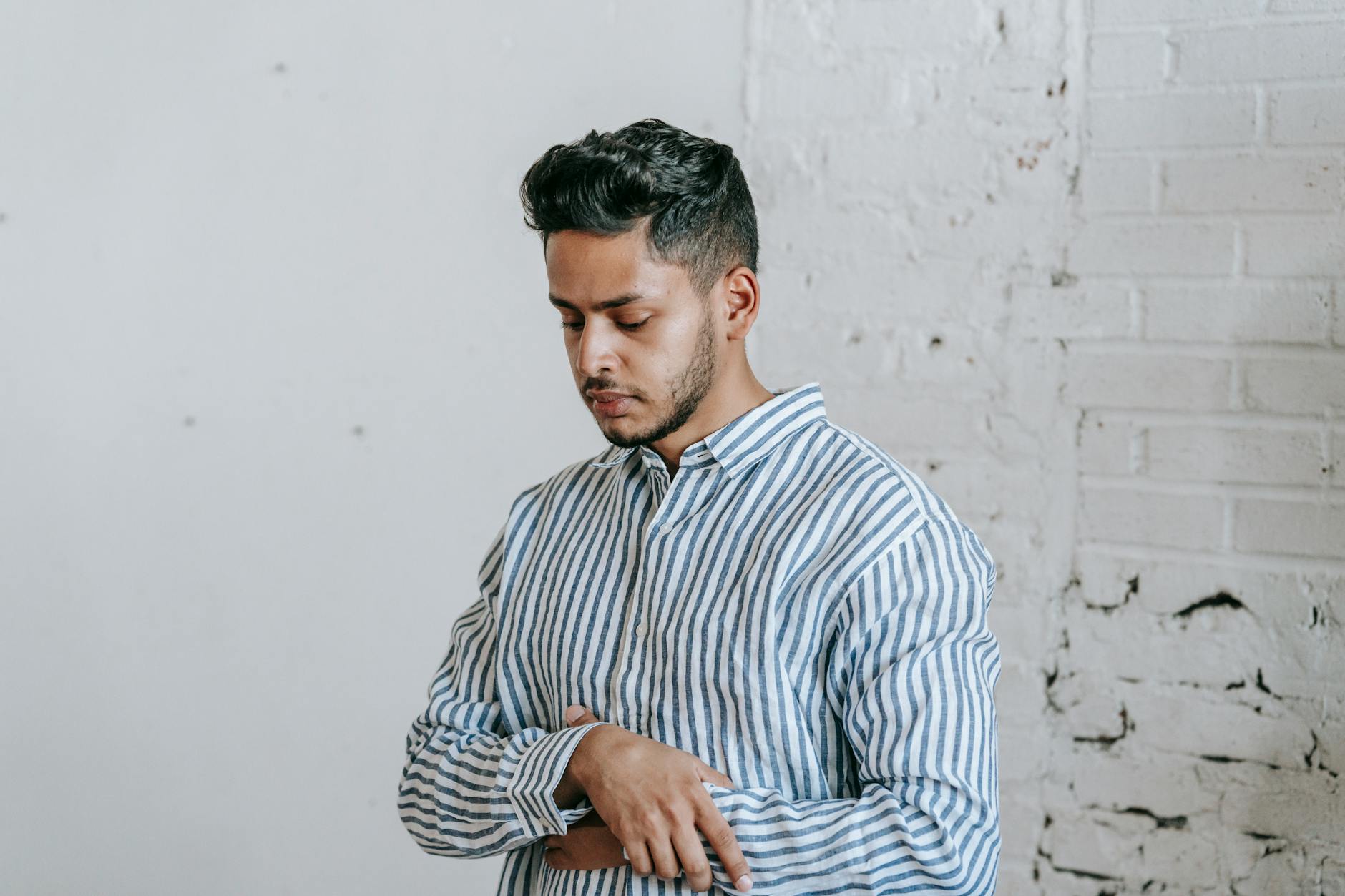 A young man standing in front of a white brick wall. He is wearing a blue and white striped button-down shirt and has a serio...