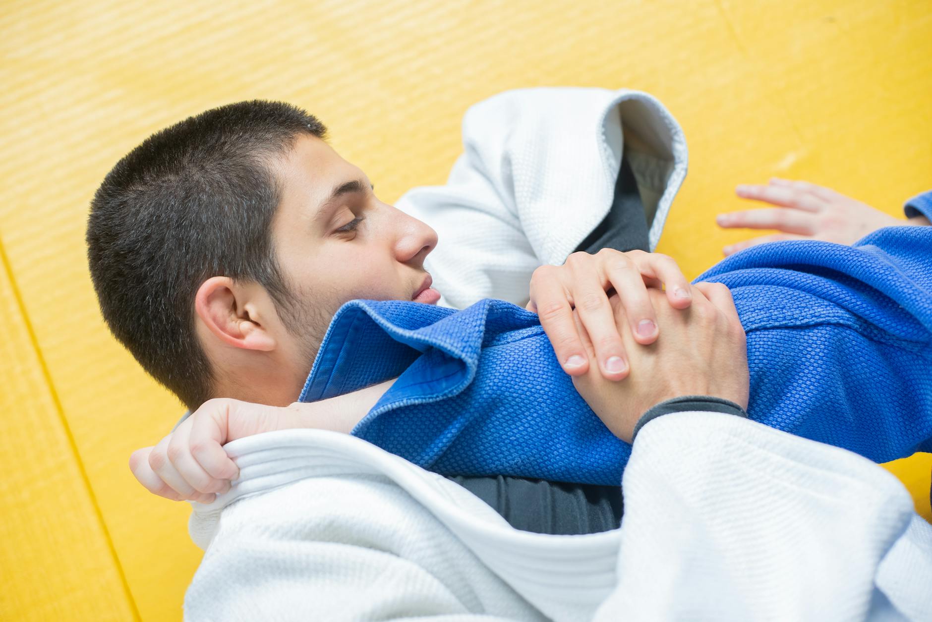 A young man in a blue judo uniform lying on his back on a yellow mat. He is wearing a white judo gi and appears to be in the ...