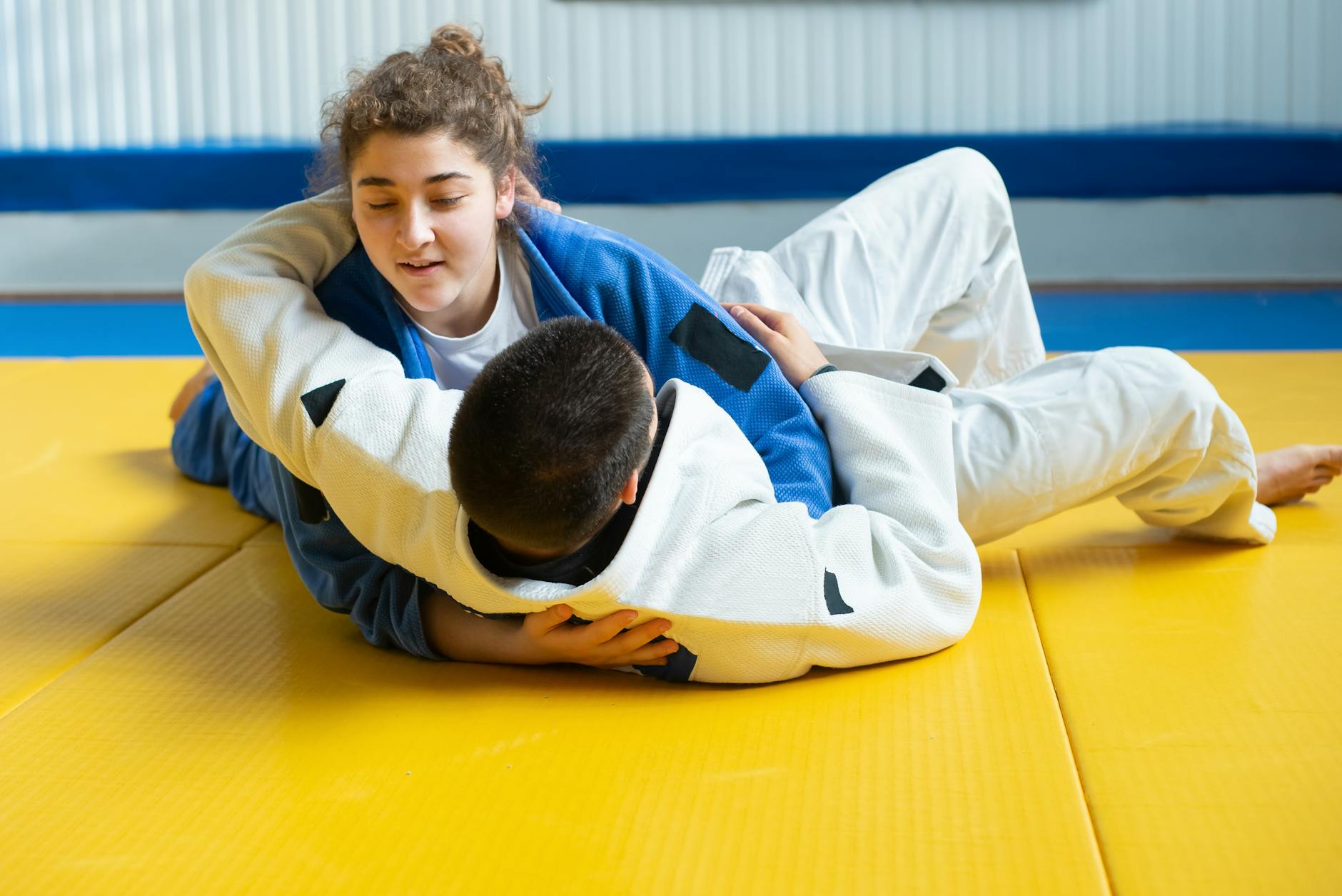 Two young people engaged in a martial arts match. The person on the left is wearing a blue and white judo uniform and is in a...