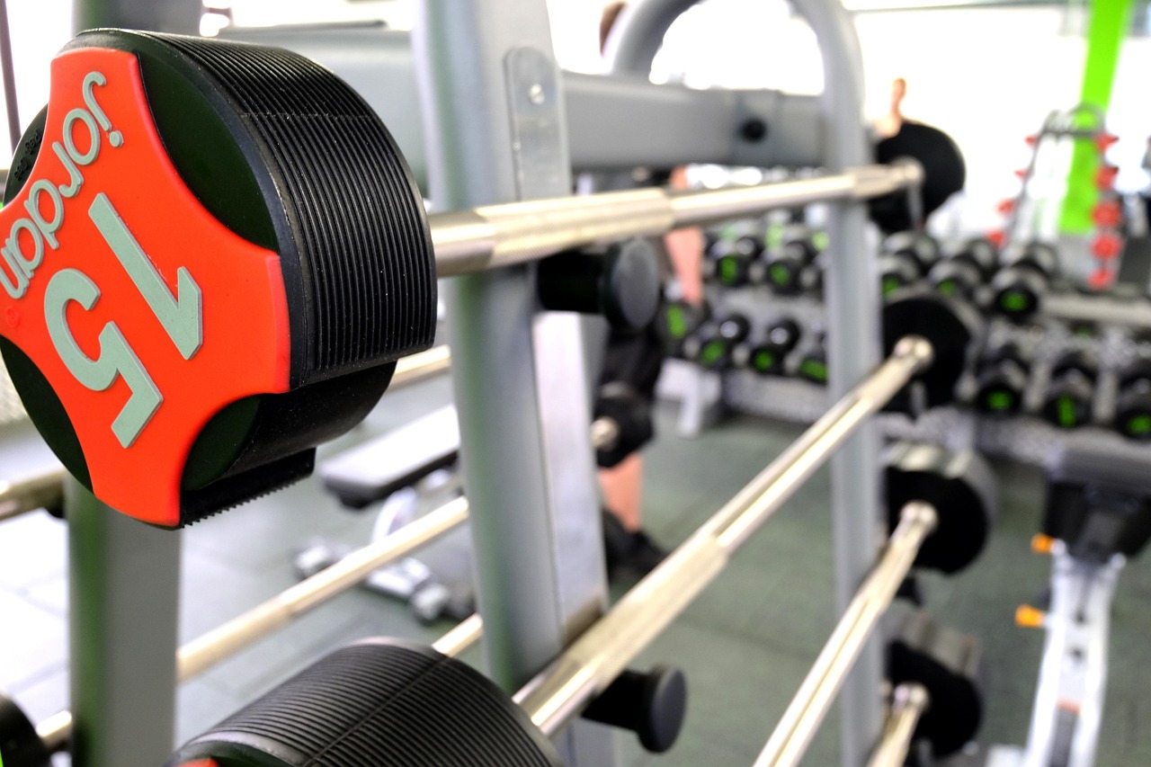 A close-up of a dumbbell rack in a gym. The rack is made of metal and has a red and black weight plate with the word "Saucony...