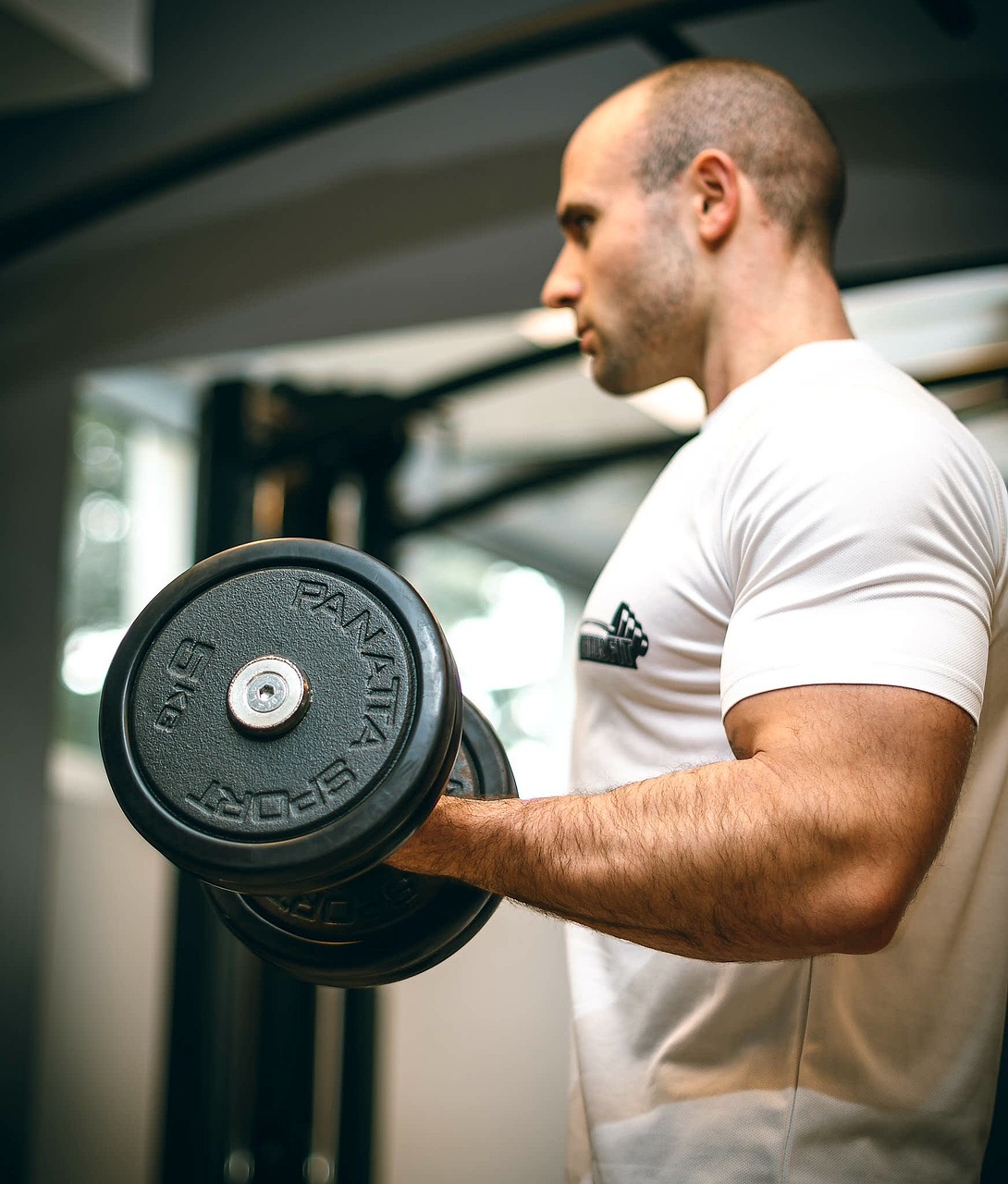 A man lifting a barbell in a gym. He is wearing a white t-shirt with a black logo on it and is standing in front of a gym equ...