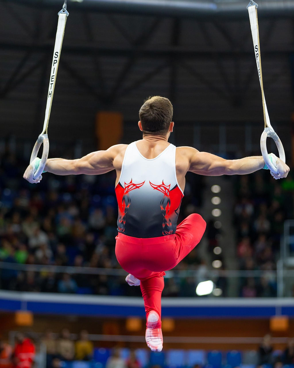 A male gymnast performing on the rings in an indoor gymnastics competition. He is wearing a red and black leotard with a red ...