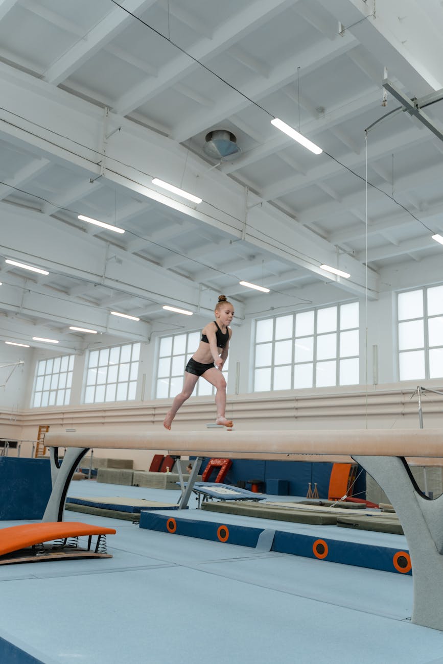 A young female gymnast performing on a balance beam in a large gymnasium. She is wearing a black leotard and black leggings a...