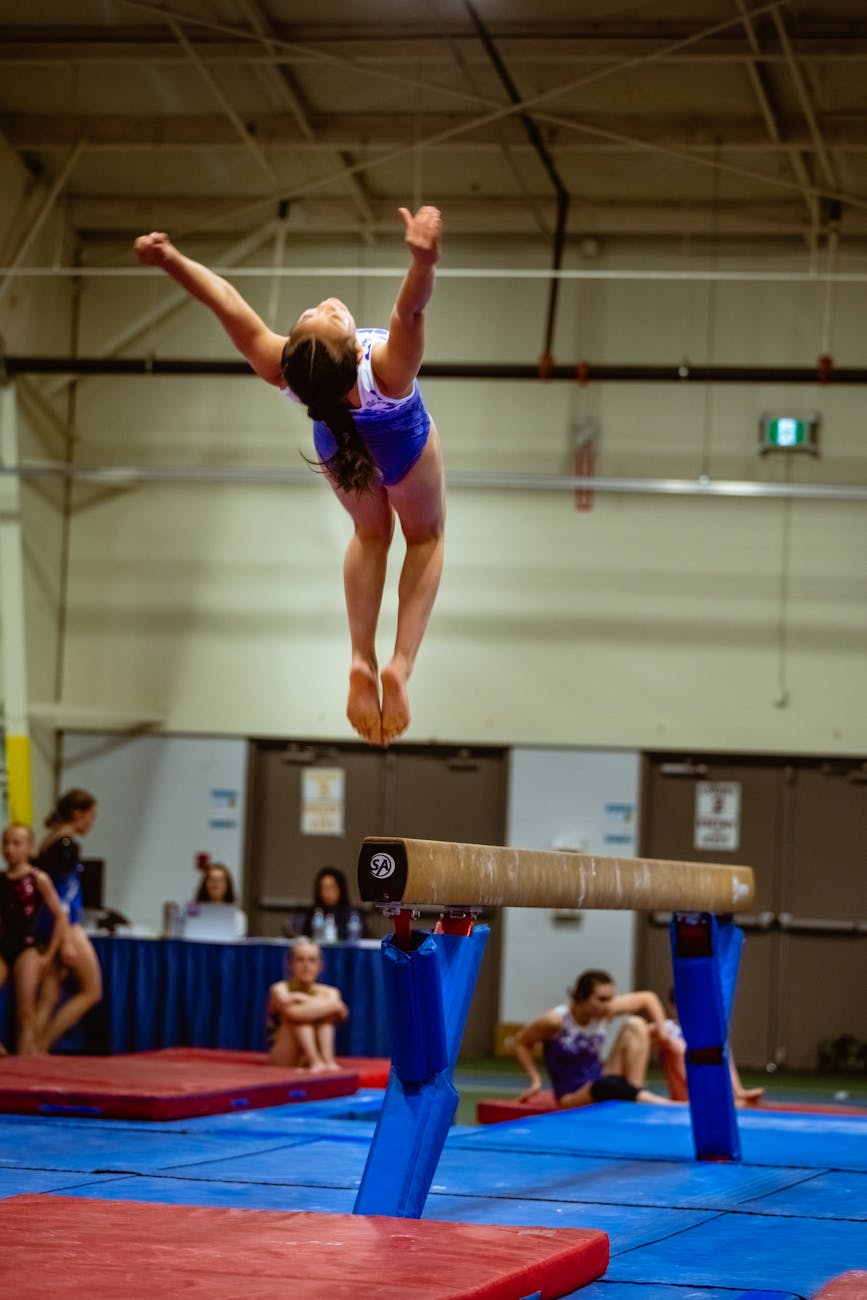 A young female gymnast performing on a balance beam in a gymnasium. She is wearing a blue leotard and is in mid-air, with her...
