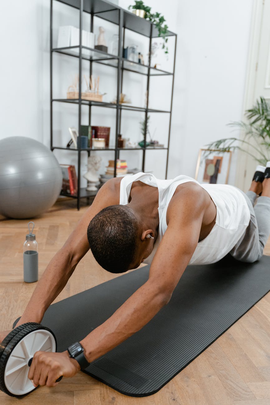 A man performing a plank exercise on a black yoga mat. He is wearing a white tank top and grey shorts. His arms are stretched out to the sides and his legs are bent at the knees. He has a black dumbbell in his right hand and a water bottle in his left hand. In the background, there is a bookshelf and a large exercise ball. The room appears to be a home gym or fitness studio.