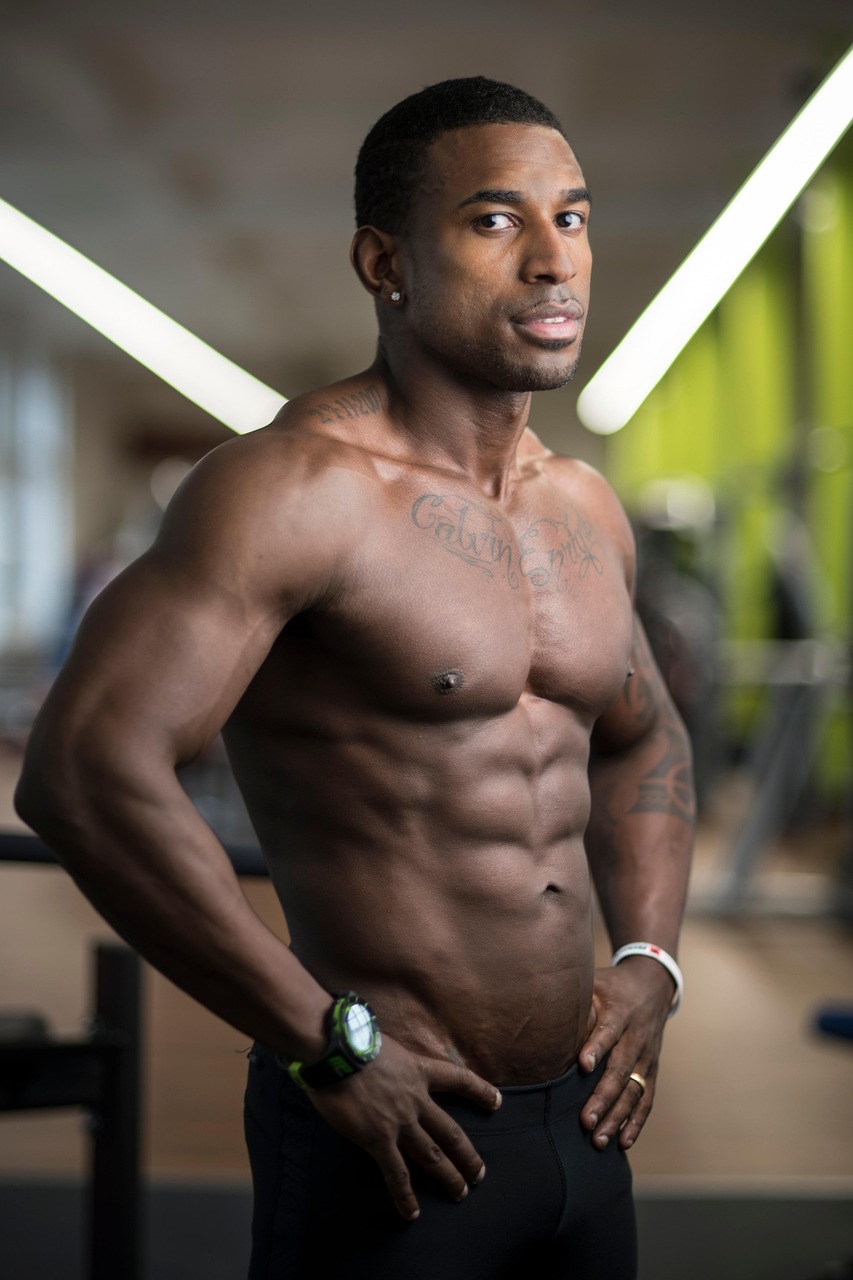 A portrait of a muscular man standing in a gym. He is shirtless and has multiple tattoos on his chest and arms. He has short ...