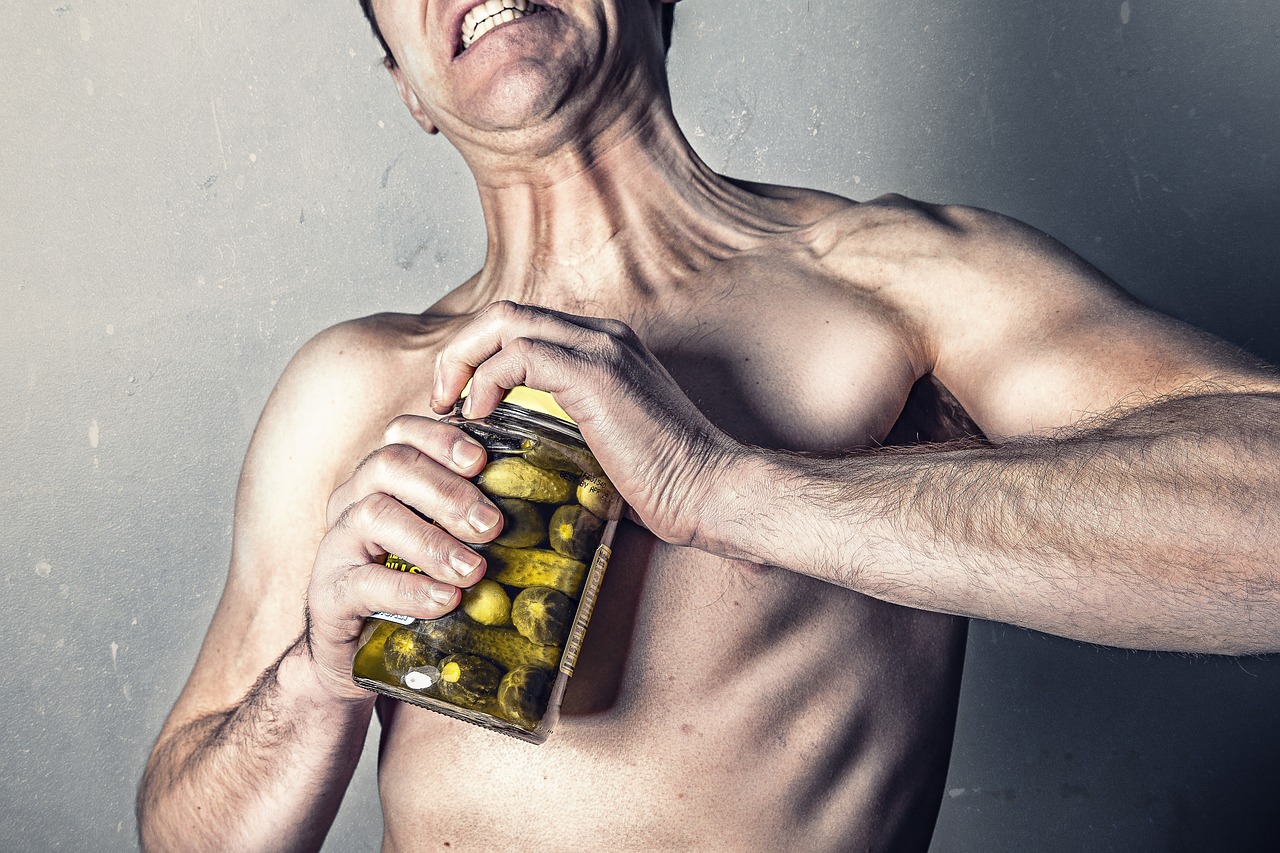 A shirtless man holding a jar of pickles in his hands. He is holding the jar with both hands and his mouth is open wide, as i...