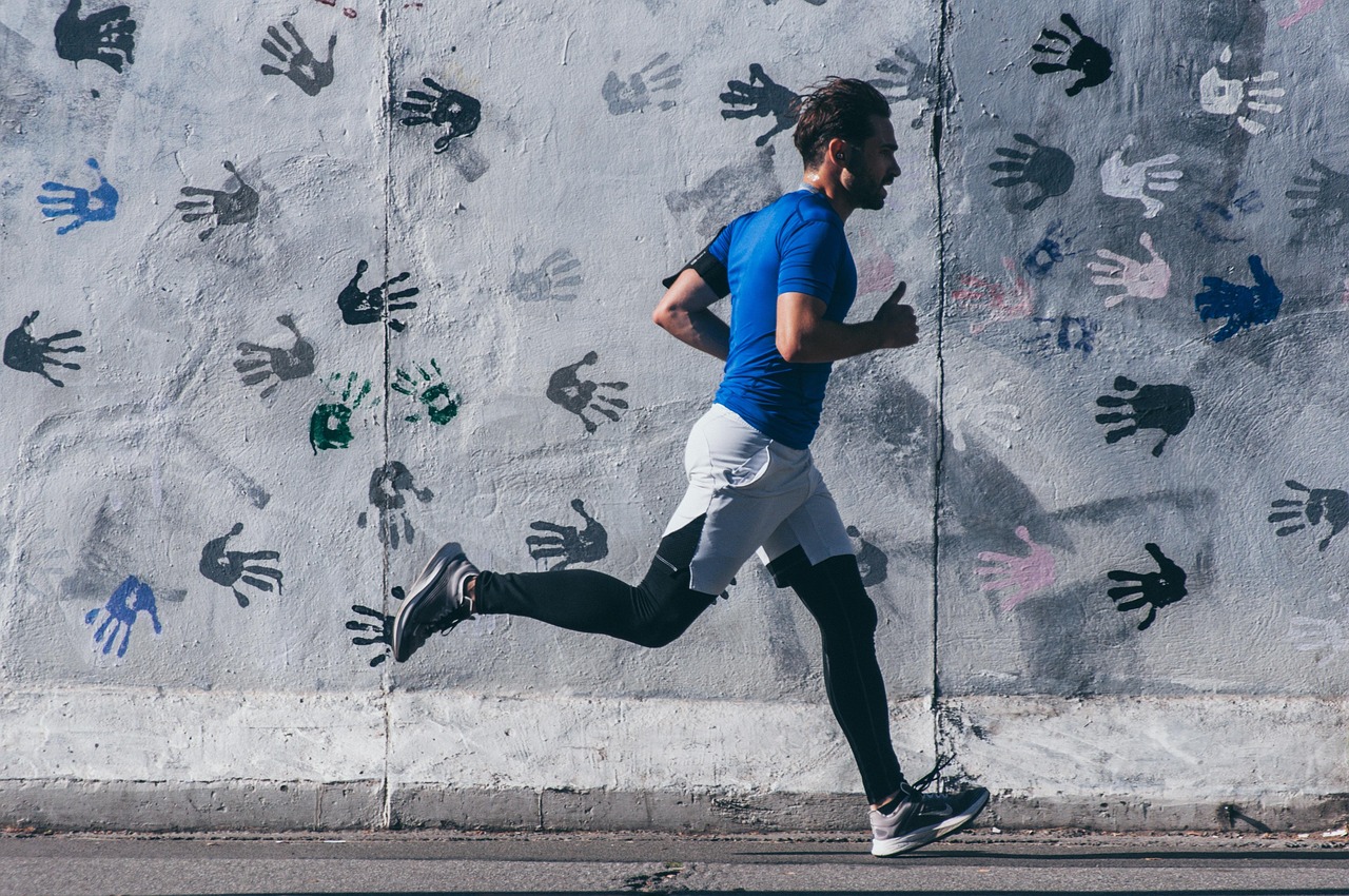 A man running on a street. He is wearing a blue t-shirt, black shorts, and white sneakers. He has a beard and is running with...