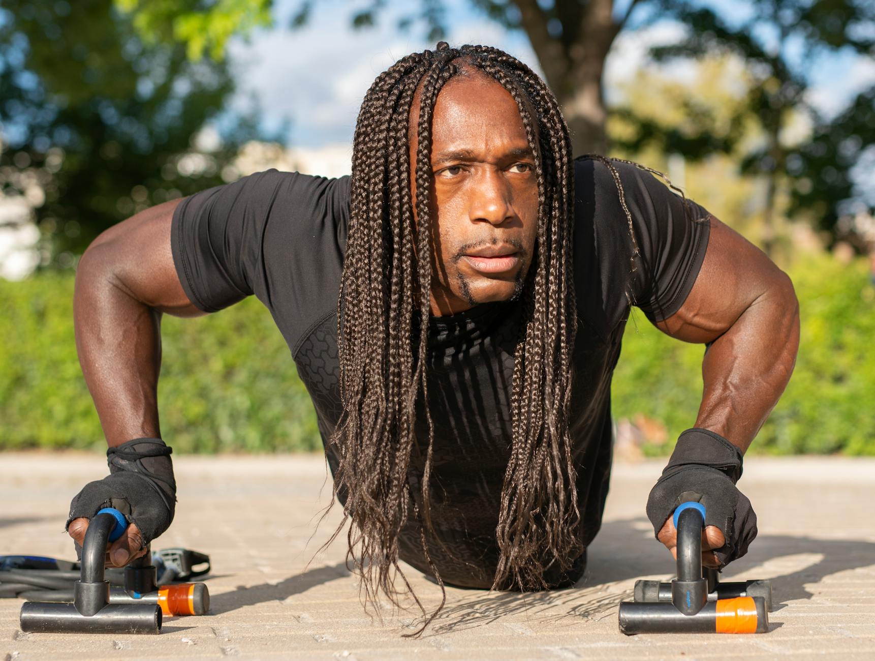 A man with long dreadlocks doing push-ups on a concrete surface. He is wearing a black t-shirt and black gloves. The man is i...