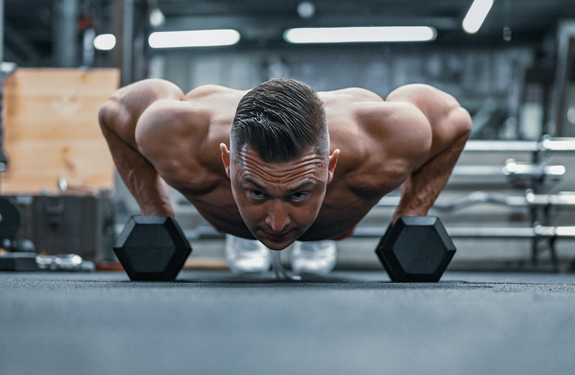 A man doing push-ups with dumbbells in a gym. He is in the middle of a plank position, with his hands and feet firmly planted...