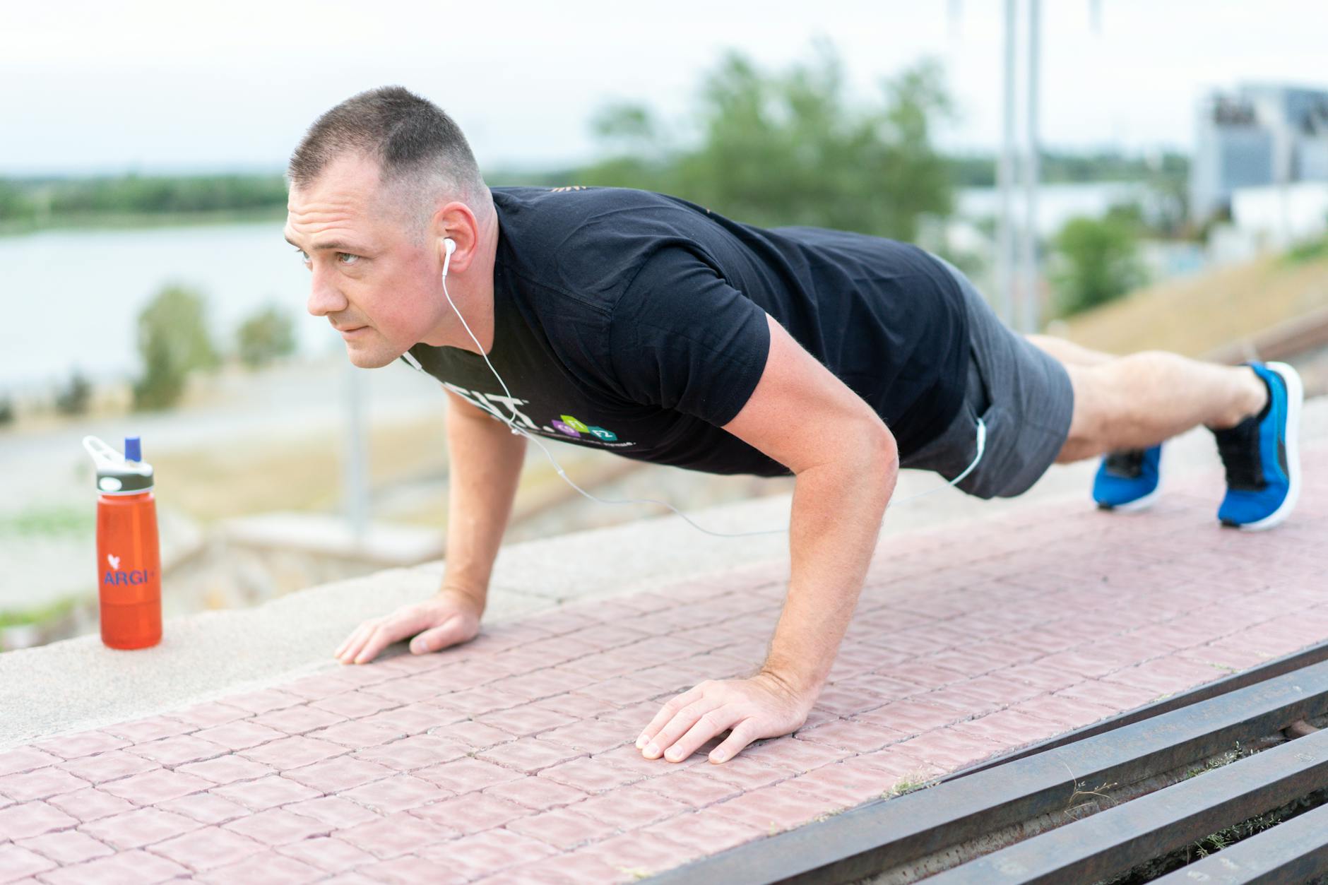 A man doing push-ups on a brick walkway. He is wearing a black t-shirt, grey shorts, and blue sneakers. He has a pair of earp...