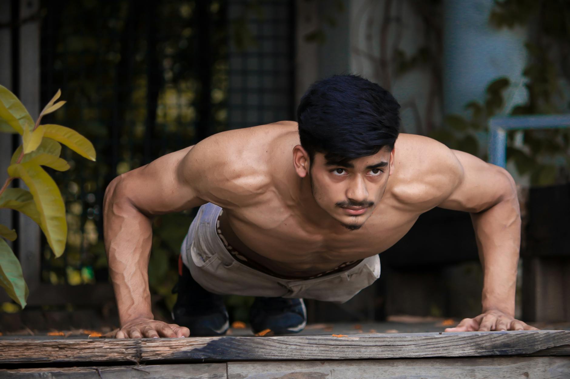A young man doing push-ups on a wooden plank. He is shirtless and wearing beige shorts. His arms are stretched out in front o...