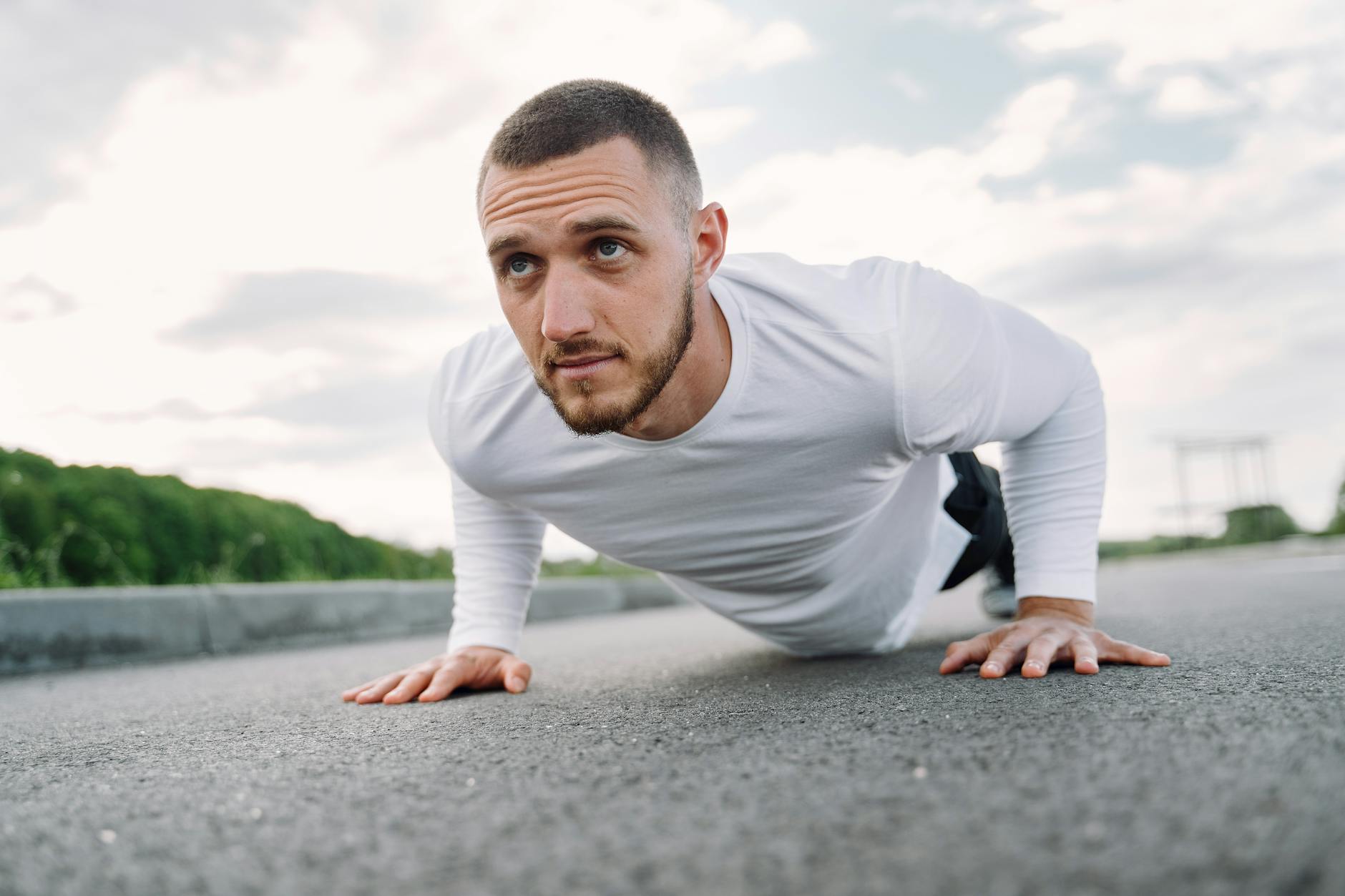 A young man doing push-ups on an empty road. He is wearing a white long-sleeved shirt and black shorts. His arms are stretche...