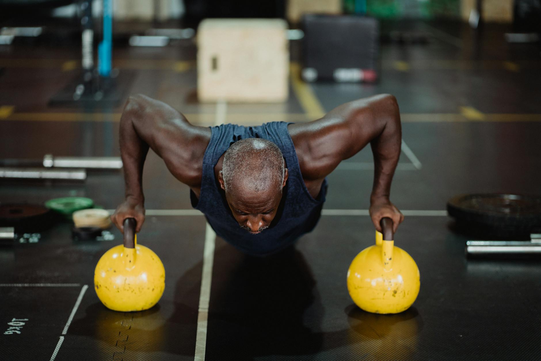 A man performing a push-up exercise with two yellow kettlebells. He is in a gym with various exercise equipment in the backgr...