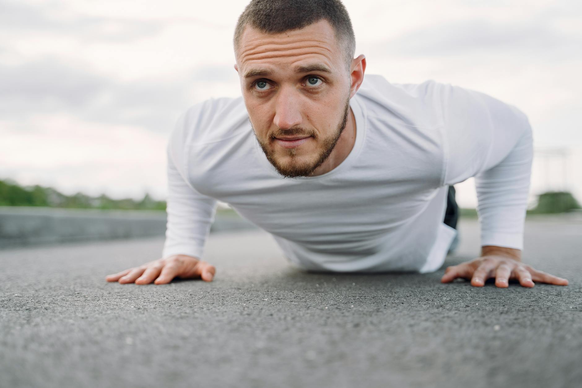 A young man doing push-ups on an empty road. He is wearing a white long-sleeved shirt and has a beard. His arms are stretched...