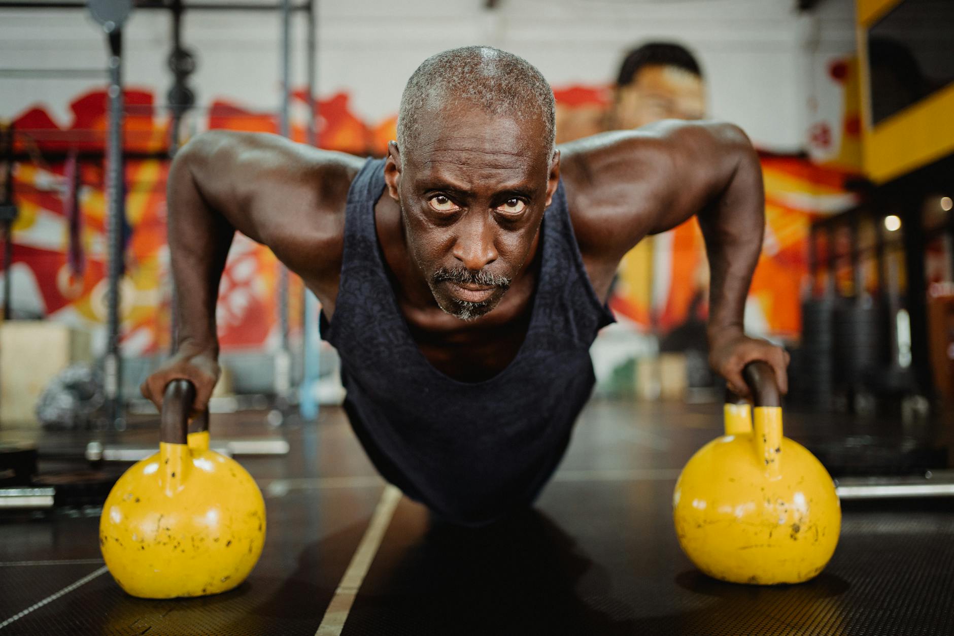 A middle-aged African-American man doing push-ups with yellow kettlebells in a gym. He is wearing a black tank top and is in ...