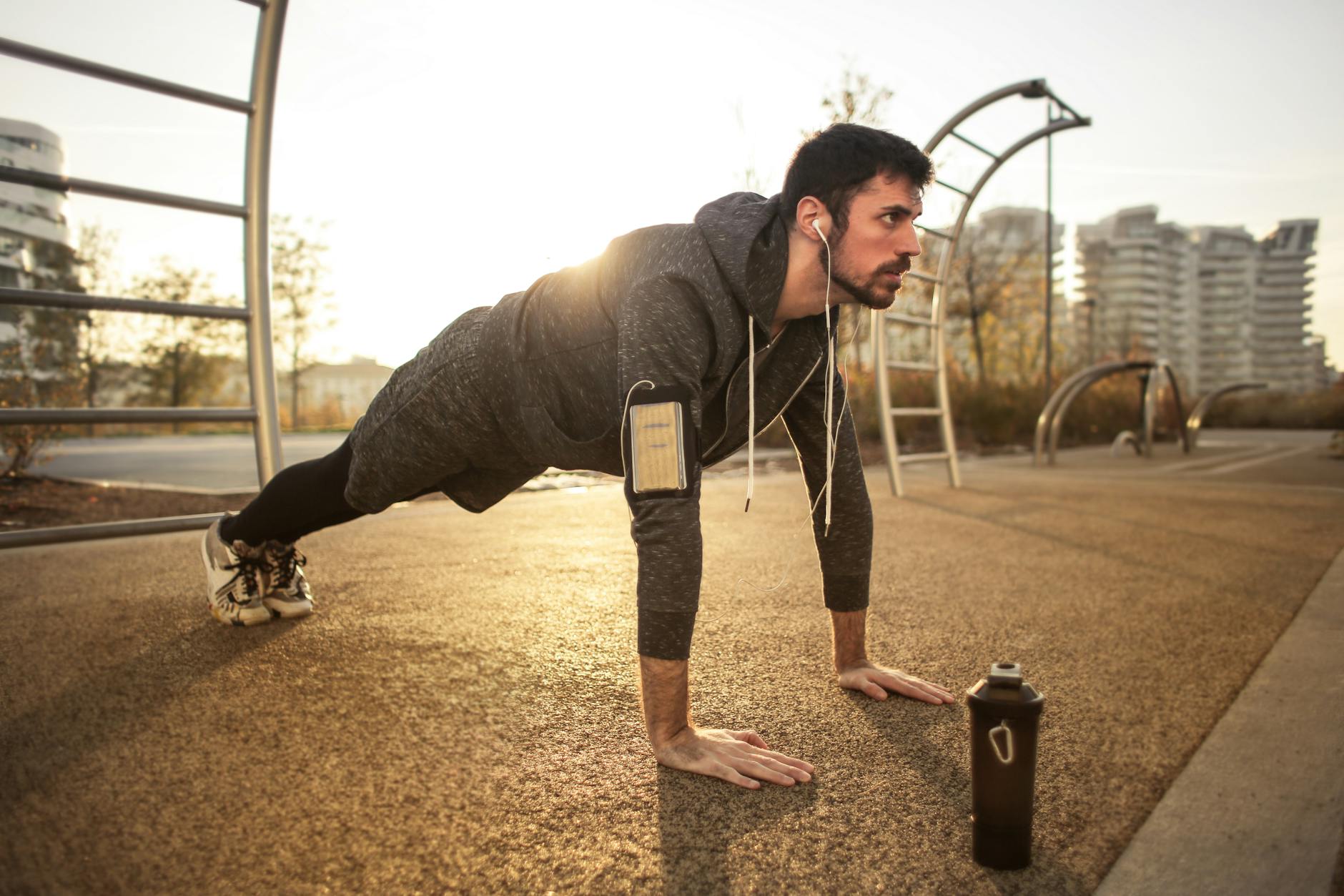 A young man doing push-ups on a concrete surface in an urban area. He is wearing a grey hoodie, black leggings, and white sne...