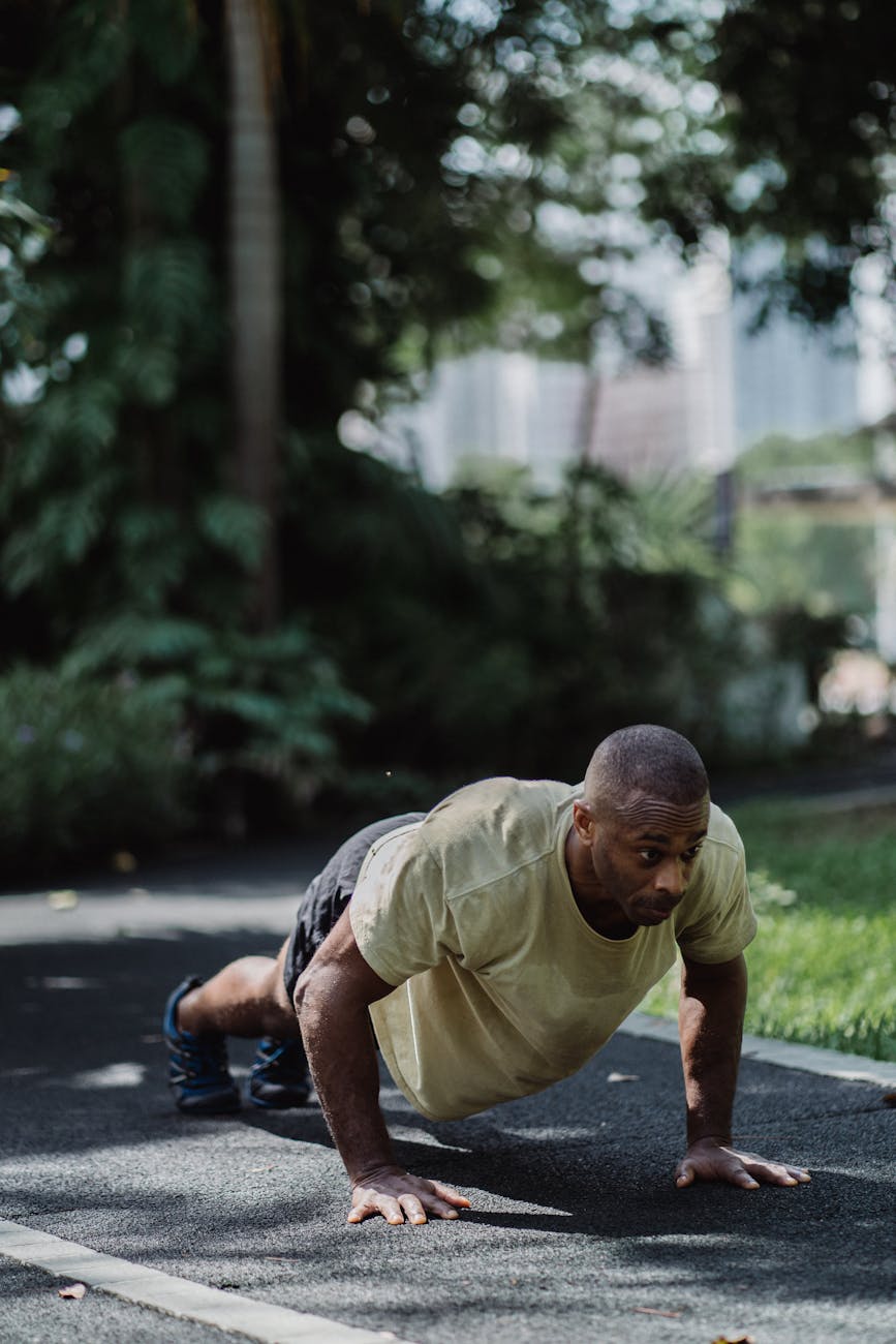 A man doing push-ups on a paved path in a park. He is wearing a yellow t-shirt and black shorts. His arms and legs are bent a...