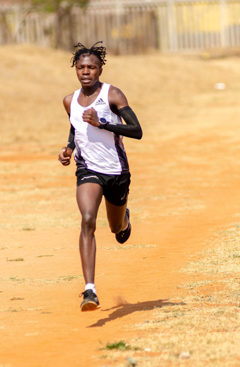A young African man running on a dirt track. He is wearing a white tank top and black shorts, and has dreadlocks on his head....