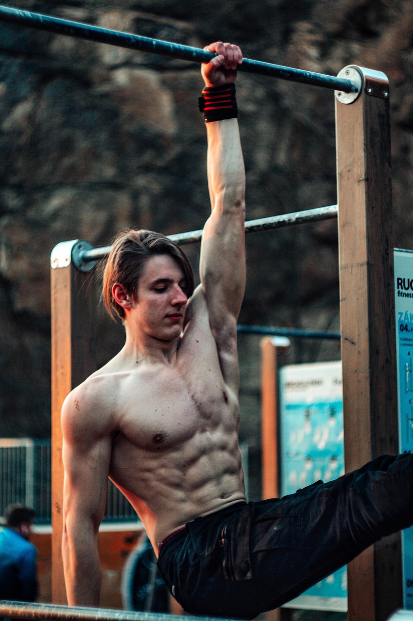 A young man performing a pull-up exercise on a horizontal bar. He is shirtless and is wearing black shorts. His arms are exte...