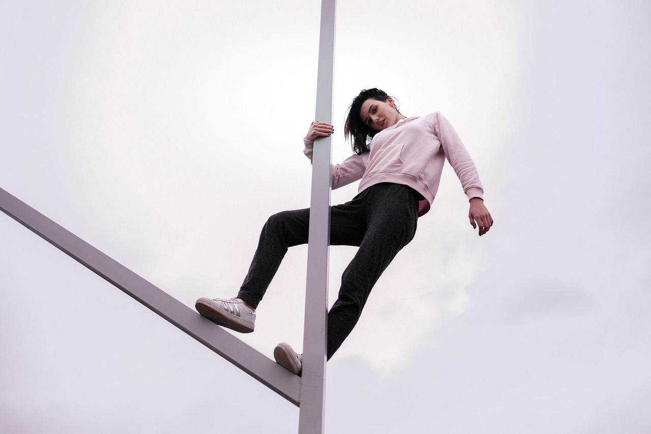 A young woman in a pink sweatshirt and black leggings climbing up a metal pole. She is holding onto the pole with both hands ...