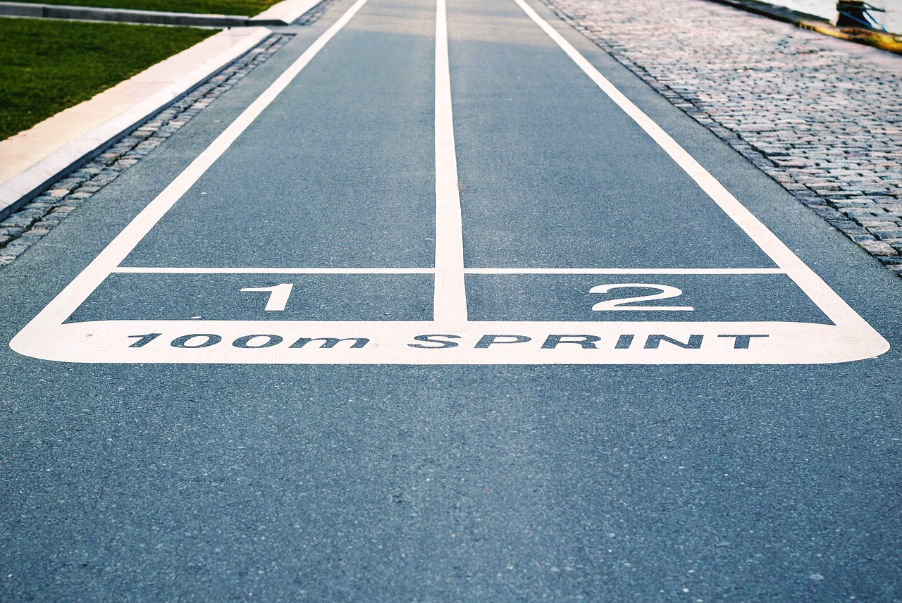 A long, straight road with a white line marking the starting point of a race. The line is divided into two sections, with the top section being the number 1 and the bottom section being a number 2. Below the line, there is a white triangle with the words "100m Sprint" written on it. The road is lined with a cobblestone pavement on one side and a grassy area on the other. The image appears to be taken from a low angle, looking down on the road.