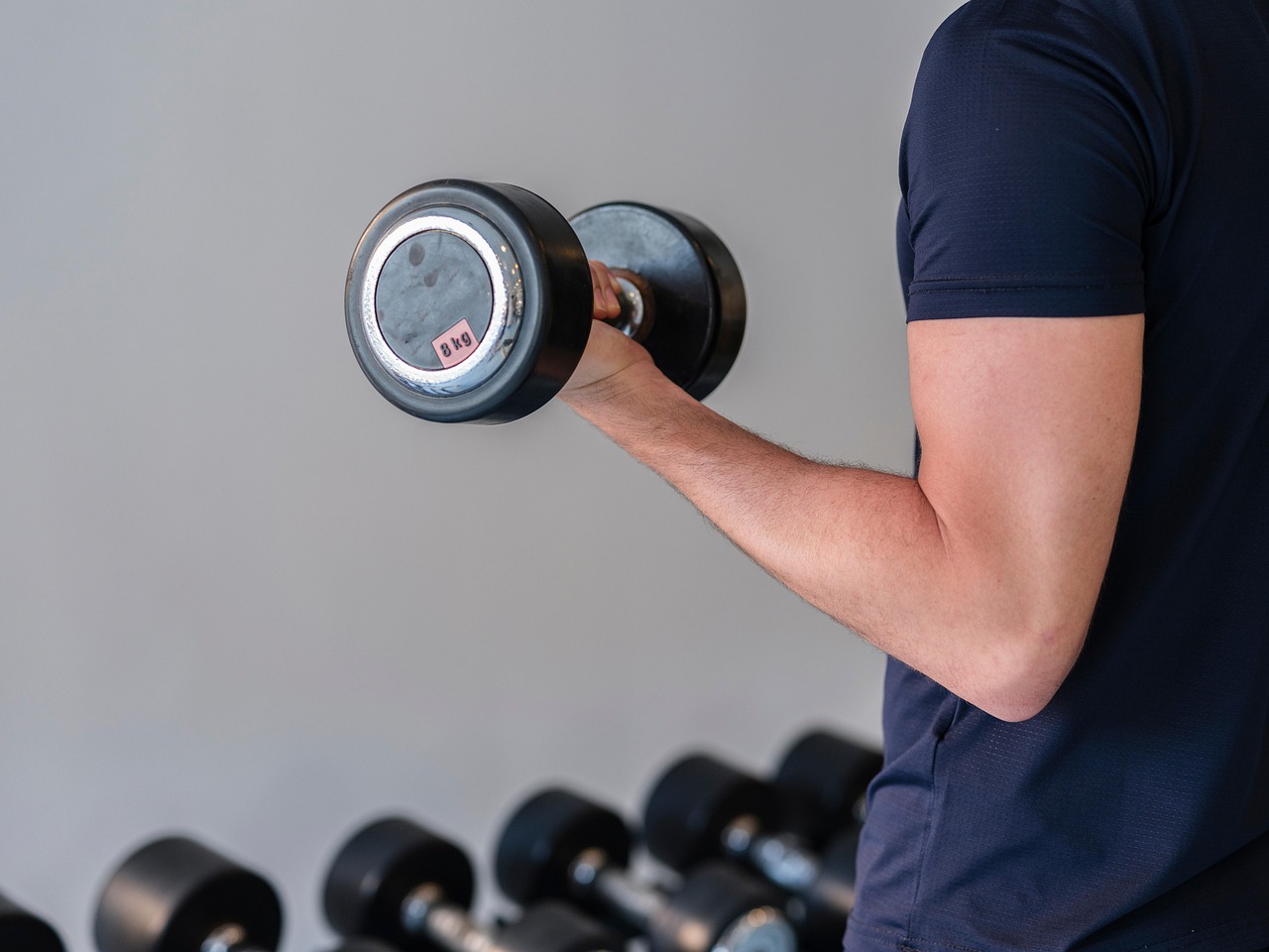 A person's arm holding a dumbbell in a gym. The person is wearing a navy blue t-shirt and is standing in front of a row of du...