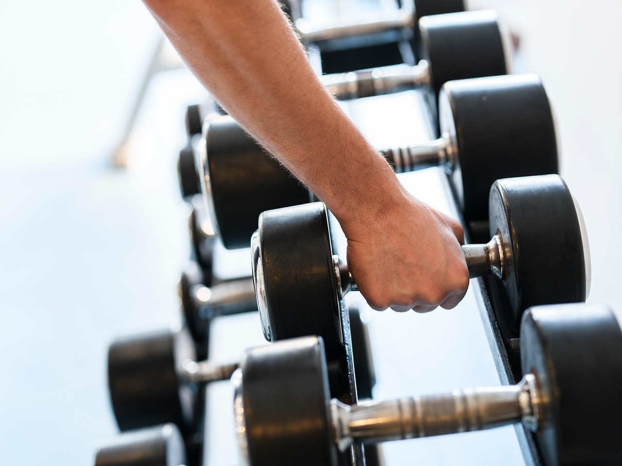 A person's hand holding a dumbbell in front of a row of dumbbells. The hand is positioned in a way that the person is lifting...