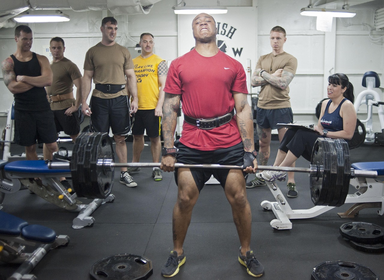 A group of people in a gym. In the center of the image, there is a man lifting a barbell with both hands. He is wearing a red...