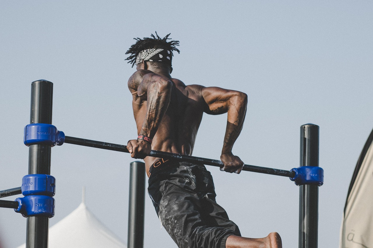 A man performing a pull-up exercise on a horizontal bar. He is wearing black shorts and a white bandana tied around his head....