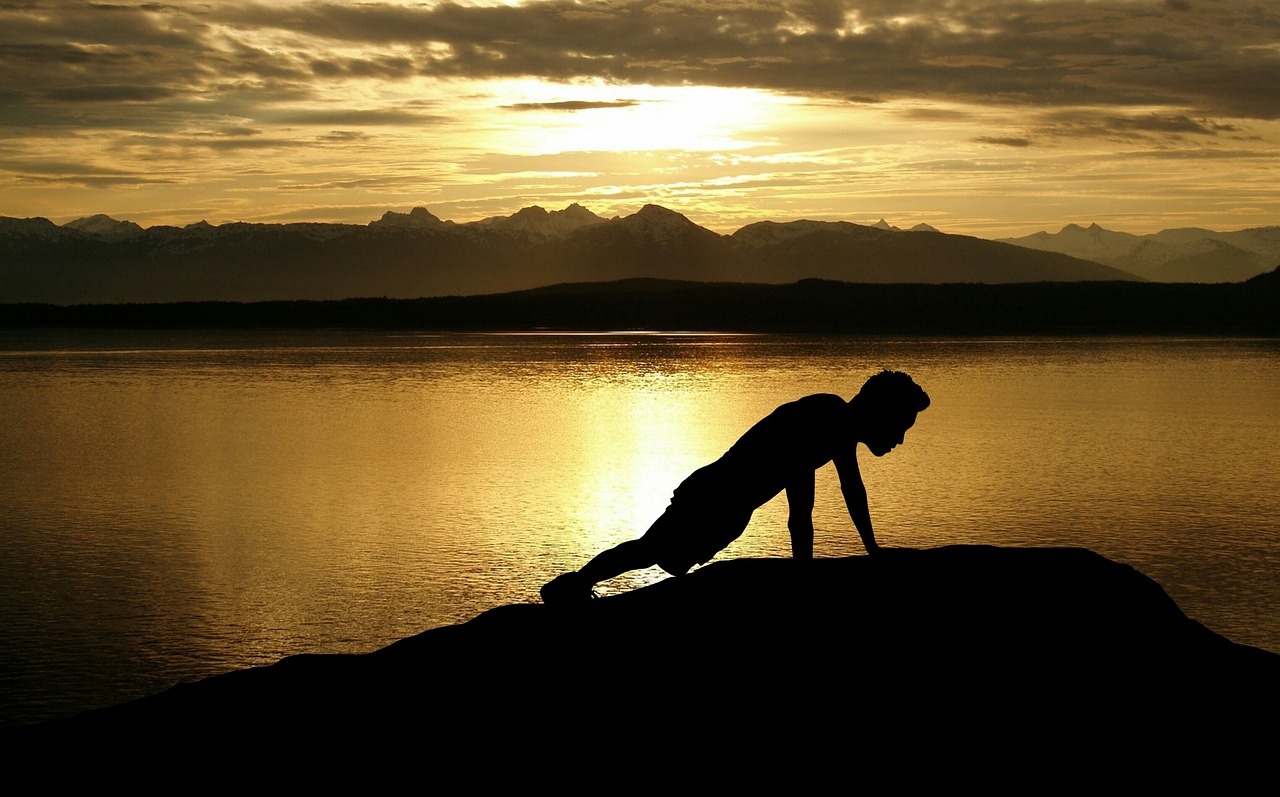 A silhouette of a person performing a yoga pose on a rock by the water. The person is in a downward facing dog pose, with the...