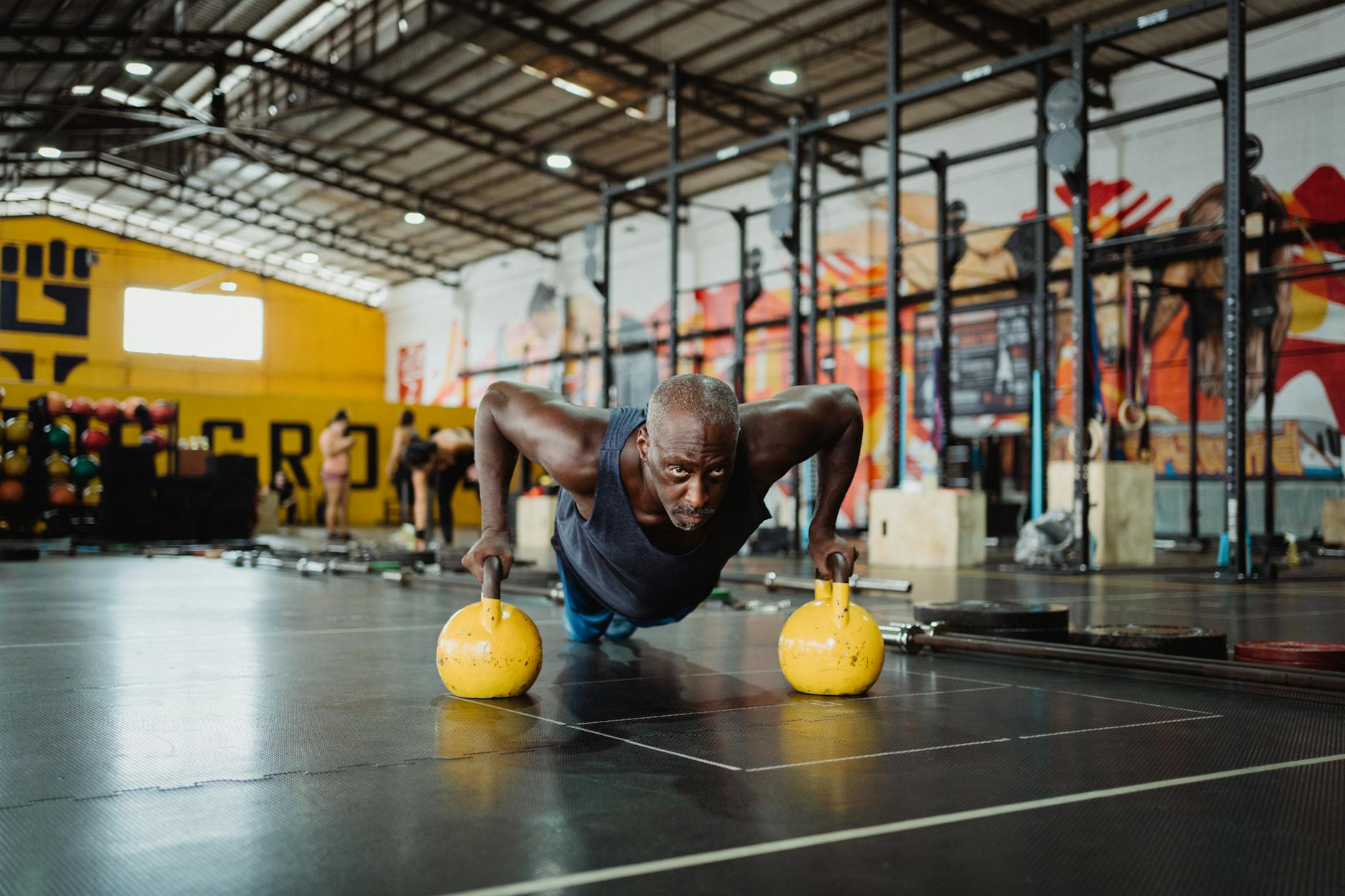 A man performing push-ups with yellow kettlebells in a gym. He is in the middle of a plank position, with his arms and legs e...