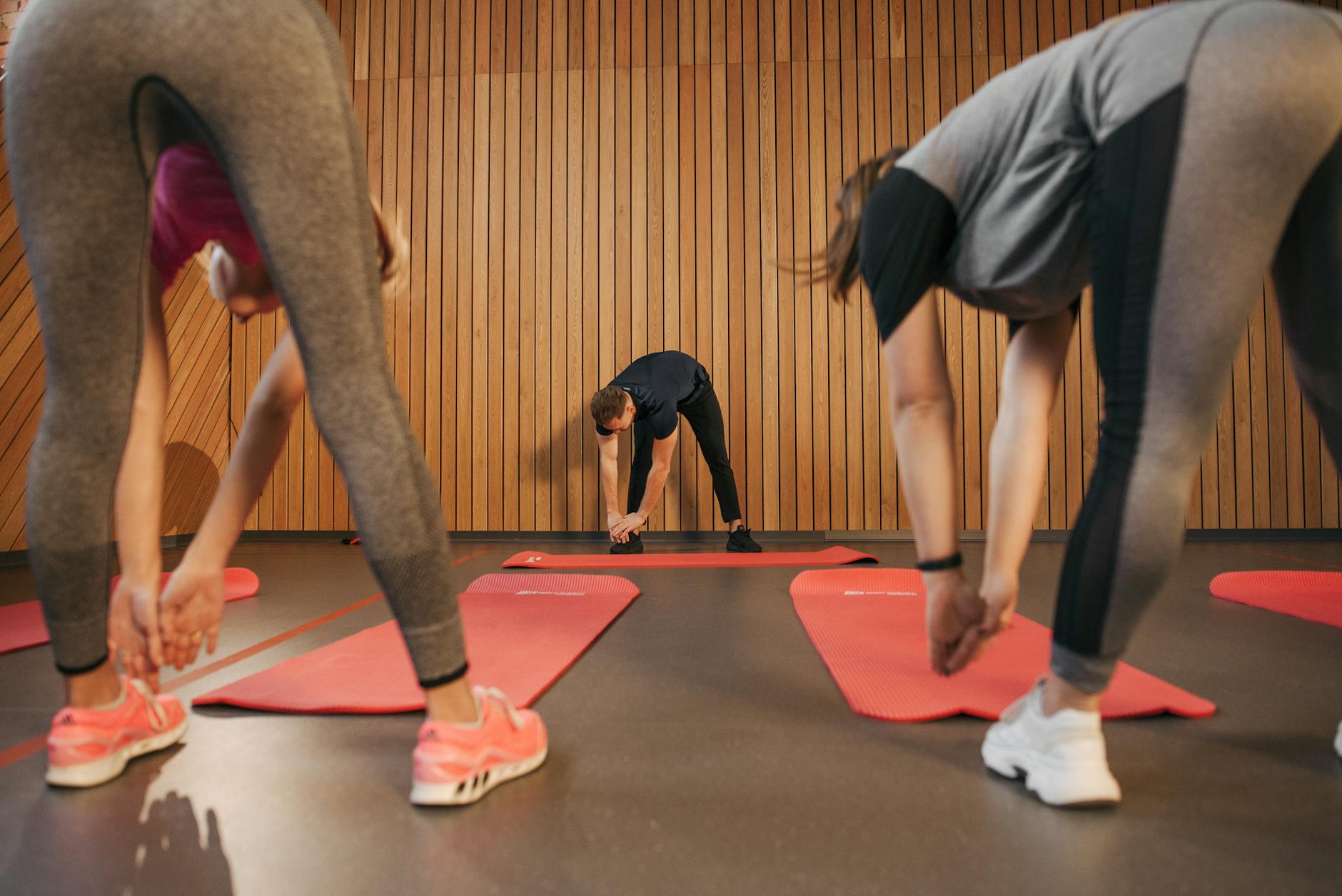 Three people in a gym, performing a yoga pose on red mats. They are all wearing workout clothes and sneakers. The person in t...