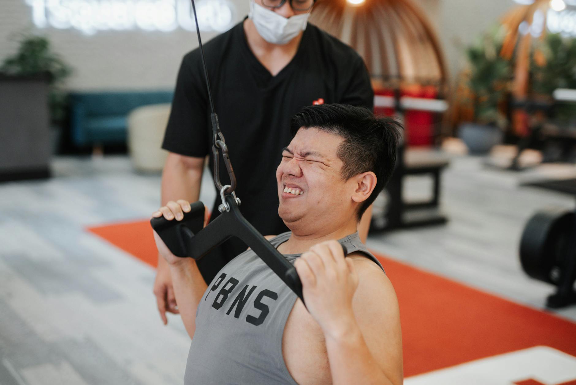 A young man in a gym, wearing a grey tank top with the word "PBS" written on it. He is holding onto a black resistance band w...