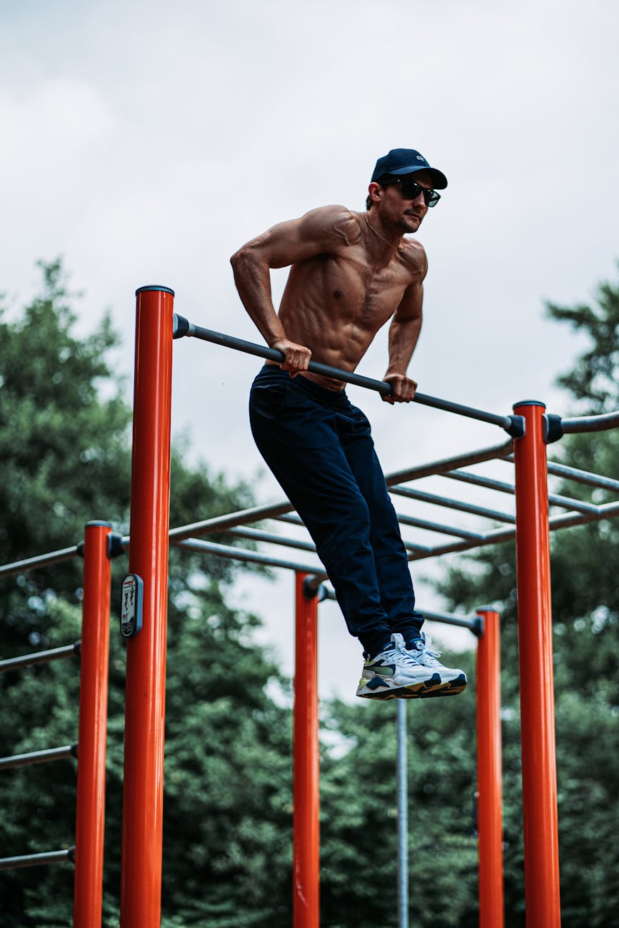 A man performing pull-ups on a horizontal bar in an outdoor gym. He is shirtless and wearing a black cap and sunglasses. The ...
