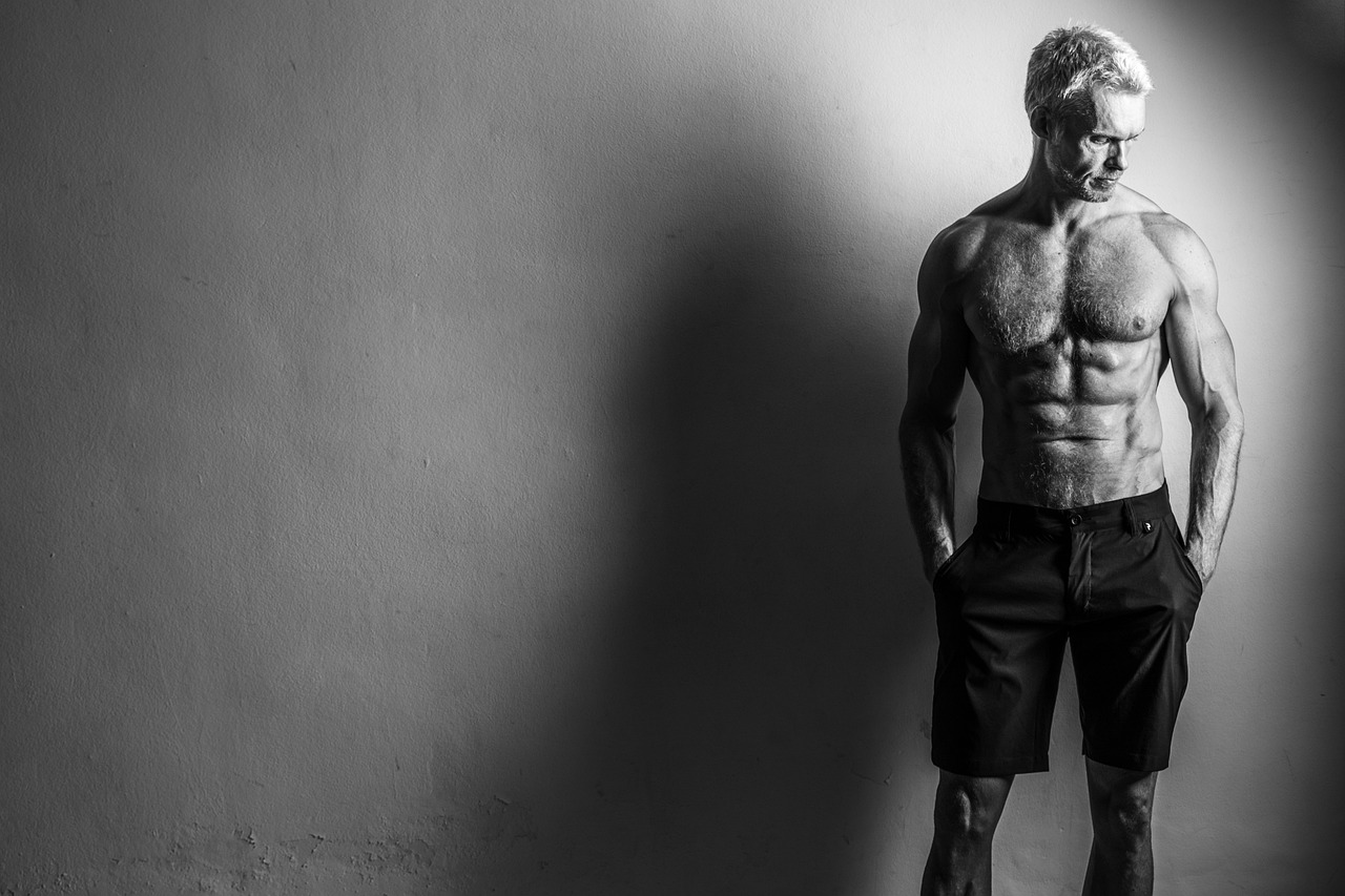 A black and white photograph of a muscular man standing in front of a plain wall. He is shirtless and wearing black shorts, w...