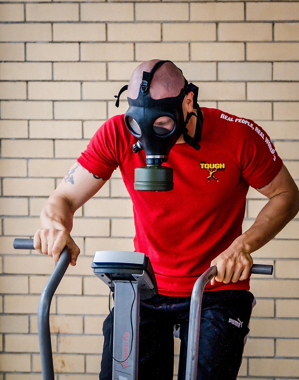 A man wearing a red t-shirt and a black gas mask. He is standing in front of a white brick wall and is holding onto the handl...
