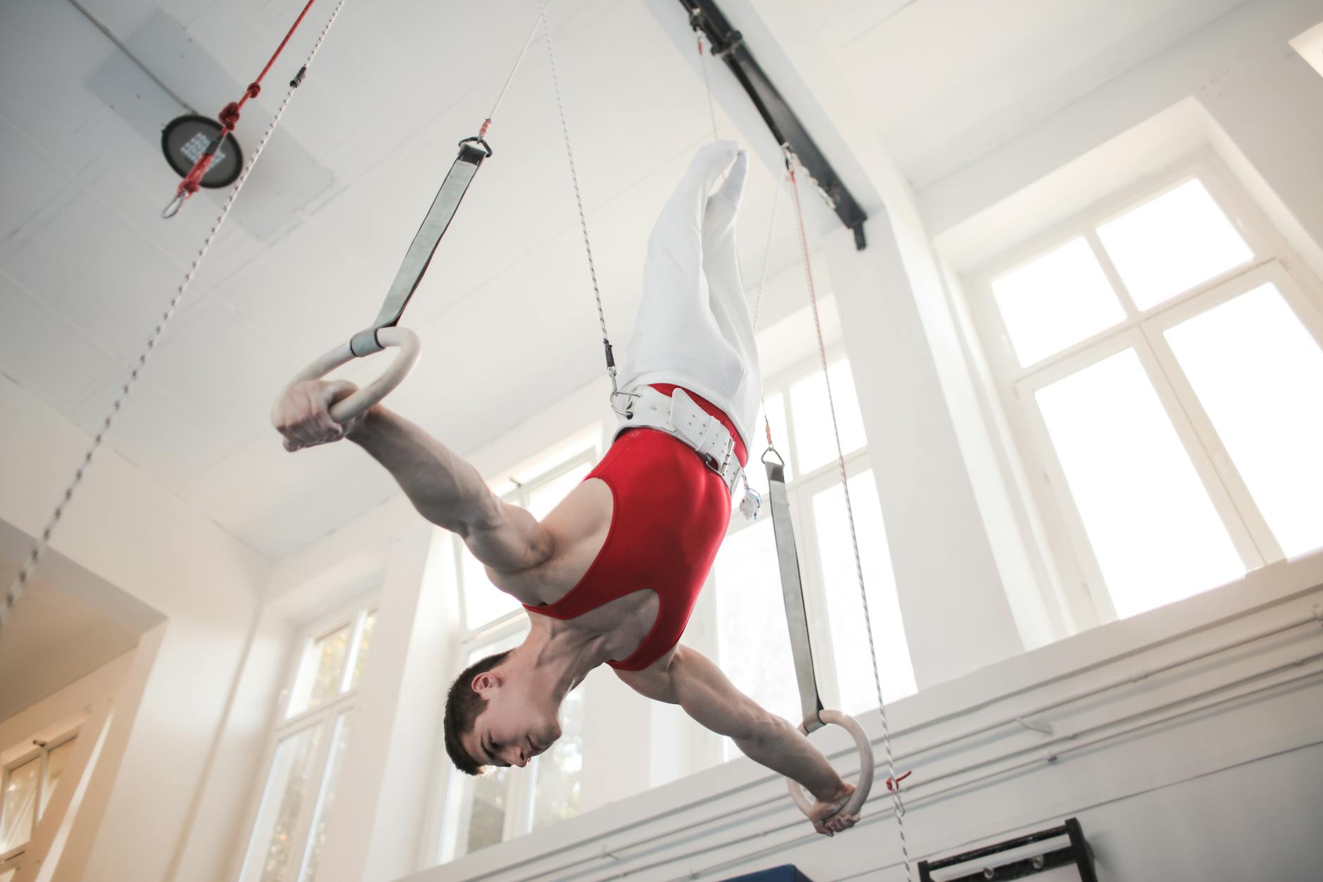 A male gymnast performing on the rings in a gymnasium. He is wearing a red leotard and white pants, and his body is in a vert...