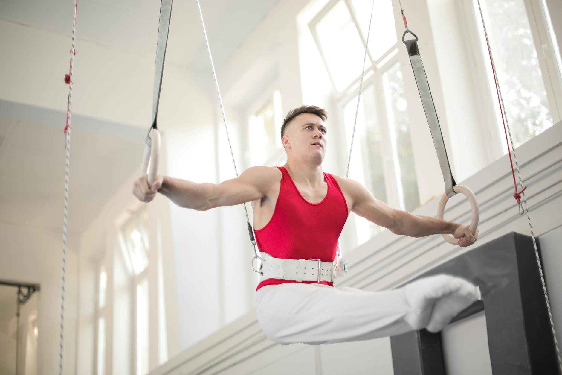 A young man performing gymnastics on rings in a gym. He is wearing a red tank top and white pants, and is holding onto the ri...