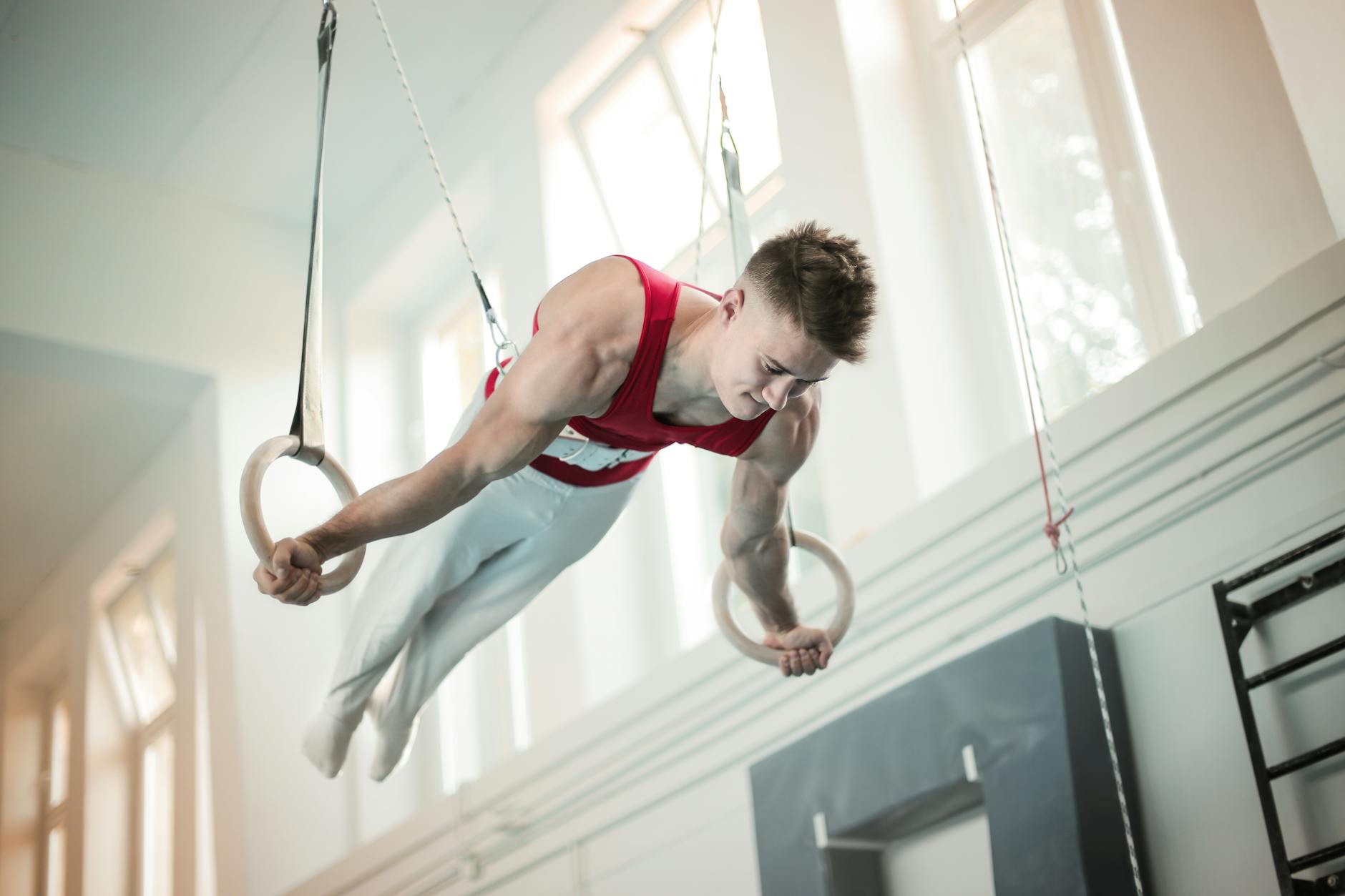 A young male gymnast performing on the rings in a gymnasium. He is wearing a red tank top and white leggings and is in mid-ai...